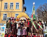 Gruppe in traditioneller Tracht vor einem Gebäude, darunter ein Maskottchen in Hasenkostüm, mit Fahne und Kunstwerken im Hintergrund.