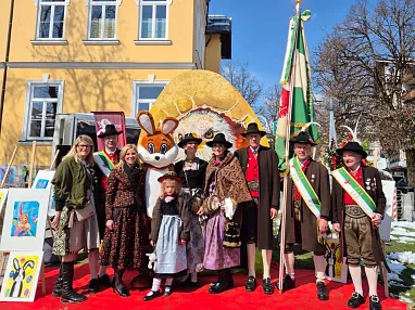 Gruppe in traditioneller Tracht vor einem Gebäude, darunter ein Maskottchen in Hasenkostüm, mit Fahne und Kunstwerken im Hintergrund.