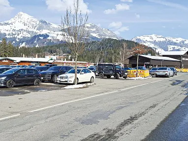 Schneebedeckte Berge hinter einem Parkplatz voller Autos an einem klaren, sonnigen Tag; vereinzelte Bäume und ein Gebäude am Rand der Straße sichtbar.