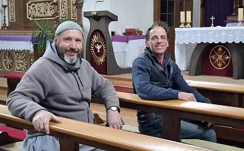 Zwei Männer sitzen in einer Kirchenbank, lächelnd. Im Hintergrund sind ein Altar und religiöse Symbole zu sehen. Die Atmosphäre wirkt freundlich und ruhig.