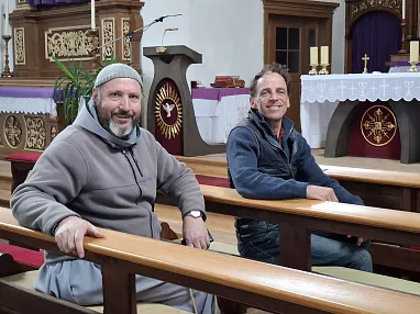 Zwei Männer sitzen in einer Kirchenbank, lächelnd. Im Hintergrund sind ein Altar und religiöse Symbole zu sehen. Die Atmosphäre wirkt freundlich und ruhig.
