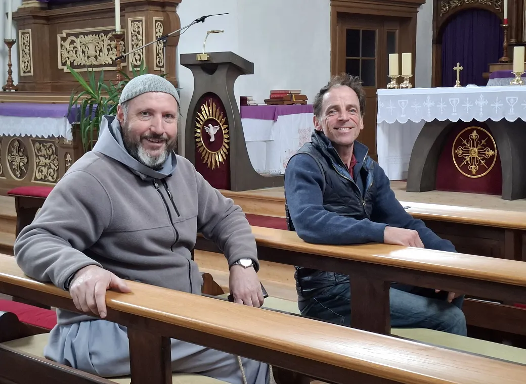 Zwei Männer sitzen in einer Kirchenbank, lächelnd. Im Hintergrund sind ein Altar und religiöse Symbole zu sehen. Die Atmosphäre wirkt freundlich und ruhig.
