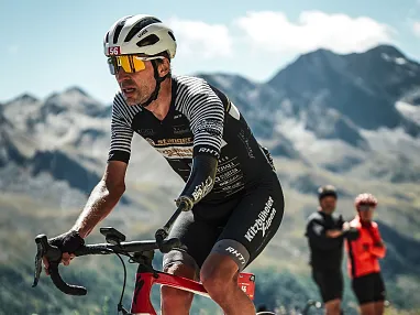 Ein Radfahrer in Rennbekleidung und Helm fährt durch eine bergige Landschaft. Im Hintergrund sind schroffe Berge und zwei Zuschauer erkennbar.