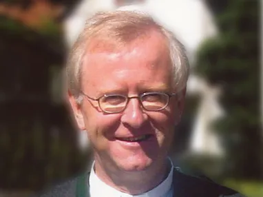 Ein älterer Mann mit Brille und hellem Haar lächelt in die Kamera. Er trägt einen Anzug mit einem weißen Hemd darunter. Der Hintergrund ist unscharf.