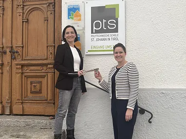 Zwei Frauen stehen lächelnd vor einem Schild der Polytechnischen Schule St. Johann in Tirol und halten symbolisch einen großen Schlüssel.