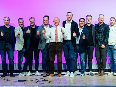 Eine Gruppe von zwölf Männern steht auf einer Bühne vor einem pink-violetten Hintergrund und zeigt Daumen hoch. Alle tragen legere Businesskleidung.