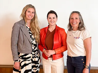 Drei Frauen stehen nebeneinander vor einer hellen Wand, lächeln und tragen verschiedene Outfits in neutralen und kräftigen Farben.