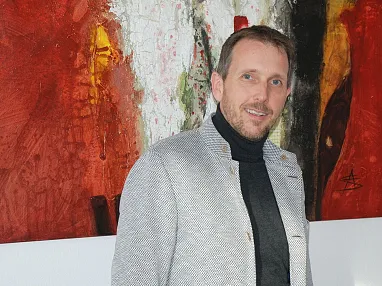 Ein Mann in einem grauen Blazer steht vor einem abstrakten Gemälde mit überwiegend roten und weißen Farben.