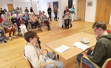 Zwei junge Männer sitzen an einem Tisch und machen eine Telefonvorführung, während ältere Personen im Hintergrund auf Stühlen und in Rollstühlen zuschauen.