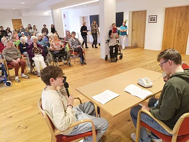 Zwei junge Männer sitzen an einem Tisch und machen eine Telefonvorführung, während ältere Personen im Hintergrund auf Stühlen und in Rollstühlen zuschauen.