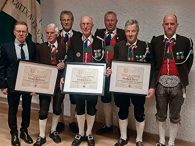 Sechs Männer in traditioneller Tracht posieren mit Urkunden vor einer Flagge. Sie stehen in einem Raum mit Holzfußboden und weißer Wand.