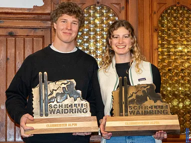 Zwei Personen halten Trophäen mit der Aufschrift "Ski Club Waidring Climbacher Alpen" vor einem Holzhintergrund. Beide lächeln in die Kamera.