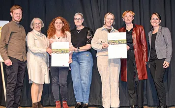 Gruppe von sieben Personen steht auf einer Bühne und hält Zertifikate, lächelt in die Kamera. Der Hintergrund ist ein dunkler Vorhang.