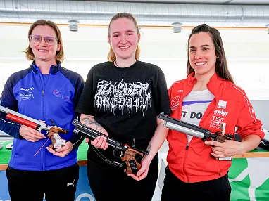 Drei lachende Frauen halten Wettkampfluftgewehre. Sie tragen Sportbekleidung und stehen vor einem Tisch mit grüner Tischdecke in einer Schießanlage.
