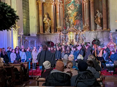 Ein Chor singt in einer reich verzierten Kirche mit goldenen Statuen und religiösen Gemälden an der Wand. Viele Menschen sitzen und hören zu.