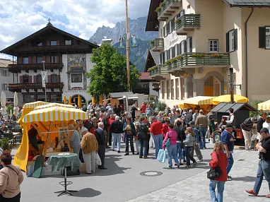 Ein belebter Marktplatz mit Ständen, Menschenmengen und historischen Gebäuden vor einem Bergpanorama an einem sonnigen Tag.