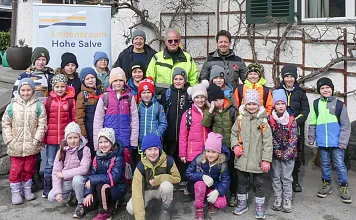 Eine Gruppe Kinder in bunter Winterkleidung steht mit Erwachsenen vor einem Haus namens "Hohe Salve". Hintergrund zeigt verschneite Berge.