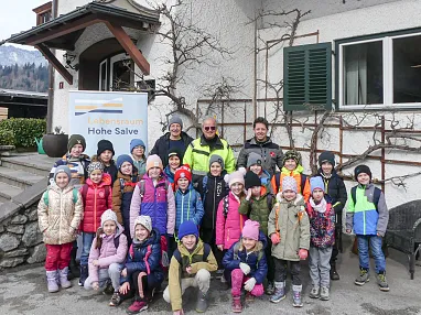 Eine Gruppe Kinder in bunter Winterkleidung steht mit Erwachsenen vor einem Haus namens "Hohe Salve". Hintergrund zeigt verschneite Berge.