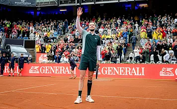 Tennisspieler auf rotem Sandplatz winkt dem Publikum zu, umgeben von einer vollen Tribüne und Bandenwerbung, Menschen im Regenmantel jubeln im Hintergrund.