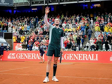 Tennisspieler auf rotem Sandplatz winkt dem Publikum zu, umgeben von einer vollen Tribüne und Bandenwerbung, Menschen im Regenmantel jubeln im Hintergrund.
