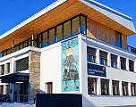 Modernes Gebäude mit großen Fenstern und einem Wandgemälde, das eine Figur zeigt, bei strahlend blauem Himmel. Ein Schild weist auf eine technische Fachschule hin.