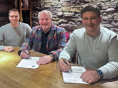 Vier Männer sitzen an einem Holztisch in einem Raum mit Steinwänden und Holzbalkendecke. Sie lächeln in die Kamera, ein Mann hält einen Stift in der Hand.