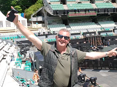 Ein Mann in Freizeitkleidung posiert fröhlich in einem leeren Stadion, dessen Sitzreihen größtenteils grün sind, mit einer aufgebauten Bühne im Hintergrund.