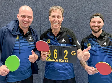 Drei Männer in Tischtennis-Shirts halten Schläger und einen Punktestand von 6:2. Sie lächeln und zeigen Daumen hoch vor einem blauen Hintergrund.