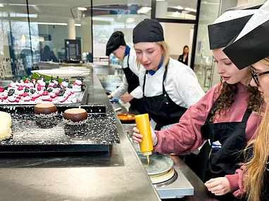 Jugendliche in Kochschürzen dekorieren Desserts in einer modernen Küche, während ein Tablett mit fertig dekorierten Backwaren daneben steht.