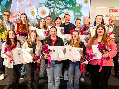 Gruppe von Menschen hält Zertifikate und Blumen, lächelnd bei einer Zeremonie. Im Hintergrund ist ein Banner mit "WKO" und "SKILLS" sichtbar.