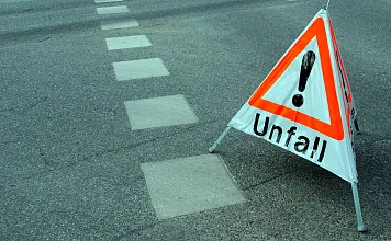 Ein Warndreieck mit der Aufschrift "Unfall" steht auf einer Straße, die Fahrbahnmarkierungen zeigt. Es weist auf einen Verkehrsunfall hin.
