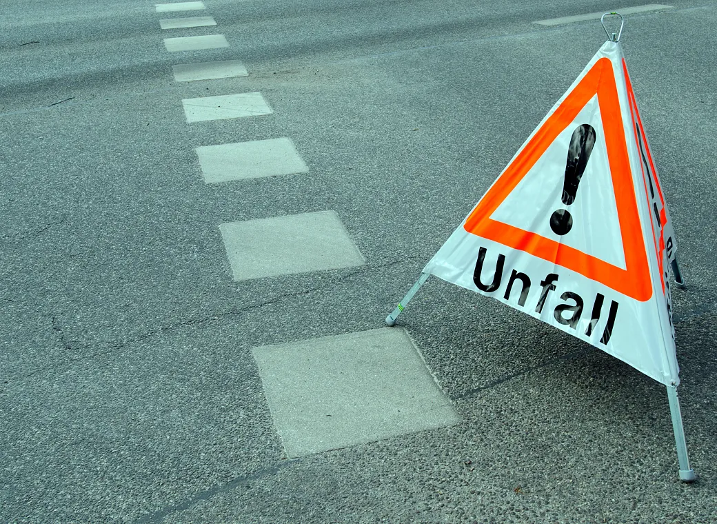 Ein Warndreieck mit der Aufschrift "Unfall" steht auf einer Straße, die Fahrbahnmarkierungen zeigt. Es weist auf einen Verkehrsunfall hin.