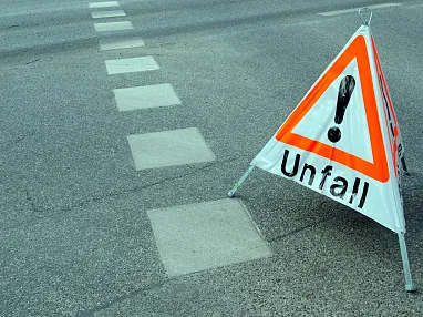 Ein Warndreieck mit der Aufschrift "Unfall" steht auf einer Straße, die Fahrbahnmarkierungen zeigt. Es weist auf einen Verkehrsunfall hin.