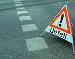 Ein Warndreieck mit der Aufschrift "Unfall" steht auf einer Straße, die Fahrbahnmarkierungen zeigt. Es weist auf einen Verkehrsunfall hin.