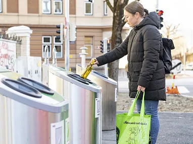 Person mit grüner Einkaufstasche wirft Flasche in öffentlichen Glasrecyclingcontainer. Im Hintergrund sind Gebäude und Straßenschilder sichtbar.