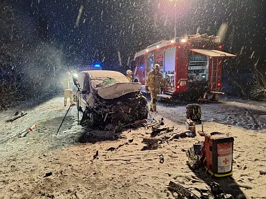 Schwer beschädigtes Auto bei Nacht auf verschneiter Straße, umgeben von Feuerwehr und Rettungskräften bei einem Unfall.