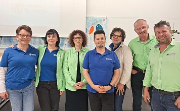 Gruppe von acht Personen in blauen und grünen Hemden posiert lächelnd vor einem Projektionsbildschirm in einem Konferenzraum.