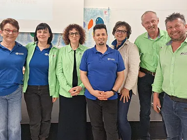 Gruppe von acht Personen in blauen und grünen Hemden posiert lächelnd vor einem Projektionsbildschirm in einem Konferenzraum.