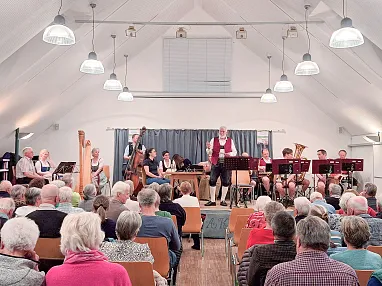 Ein traditionelles Musikensemble spielt auf einer Bühne in einem Saal mit Publikum. Die Musiker tragen Trachten und es herrscht eine freundliche Atmosphäre.