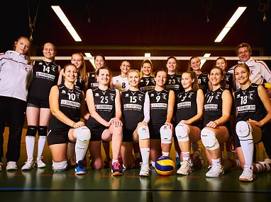 Eine Volleyballmannschaft steht in der Sporthalle, die Spielerinnen tragen schwarze Trikots mit weißen Nummern und posieren vor dem Netz für ein Teamfoto.