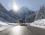 Ein gelber Bus fährt auf einer verschneiten Gebirgsstraße bei Sonnenschein. Im Hintergrund sind schneebedeckte Bäume und Berge zu sehen.