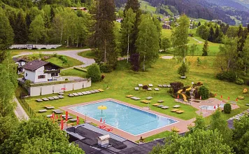 Luftaufnahme eines Freibads umgeben von grünen Wiesen und Wäldern, mit Bergen im Hintergrund. Sonnenliegen und Sonnenschirme sind am Pool verteilt.