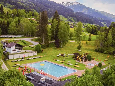 Luftaufnahme eines Freibads umgeben von grünen Wiesen und Wäldern, mit Bergen im Hintergrund. Sonnenliegen und Sonnenschirme sind am Pool verteilt.