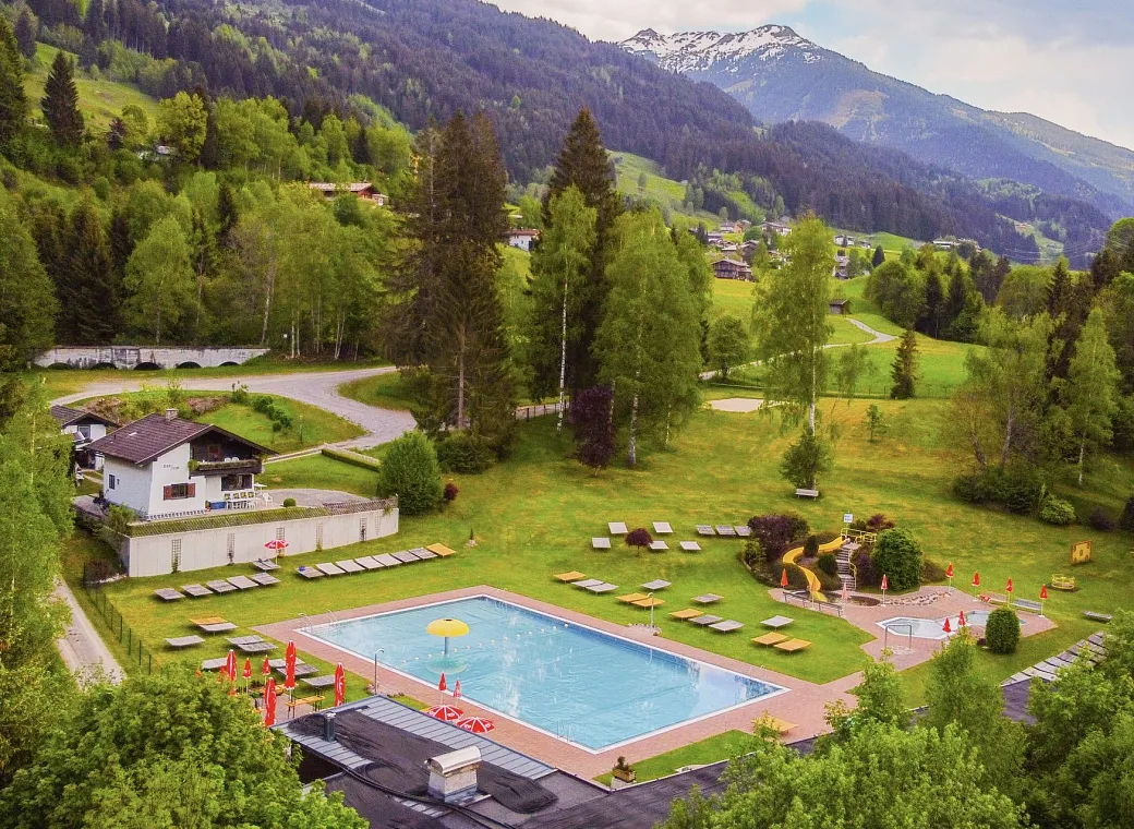 Luftaufnahme eines Freibads umgeben von grünen Wiesen und Wäldern, mit Bergen im Hintergrund. Sonnenliegen und Sonnenschirme sind am Pool verteilt.