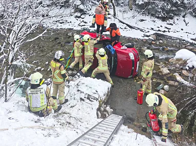 Feuerwehrleute retten Insassen aus einem Auto, das in einem verschneiten Bach liegt. Einsatzkräfte in Schutzkleidung leisten Erste Hilfe.