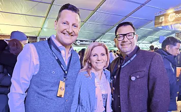 Drei lächelnde Personen in traditioneller Tracht stehen unter einem Zelt mit Beleuchtung. Die Umgebung ist belebt und festlich.