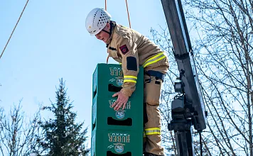 Ein Feuerwehrmann in Schutzkleidung und Helm stapelt Bierkisten draußen. Ein Kran ist im Hintergrund sichtbar. Es ist ein klarer, sonniger Tag.