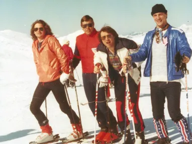Vier Männer in Ski-Outfits stehen auf einer verschneiten Piste. Sie lächeln in die Kamera und tragen bunte Jacken. Der Himmel ist klar und sonnig.
