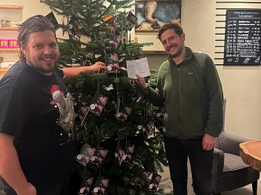 Zwei Männer stehen lächelnd neben einem festlich geschmückten Weihnachtsbaum im Innenbereich, der mit kleinen Geschenken und Lichtern dekoriert ist.
