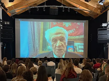 Eine große Gruppe von Menschen sitzt in einem Raum und schaut auf eine Leinwand, auf der ein älterer Mann mit weißem Haar per Videoübertragung zu sehen ist.